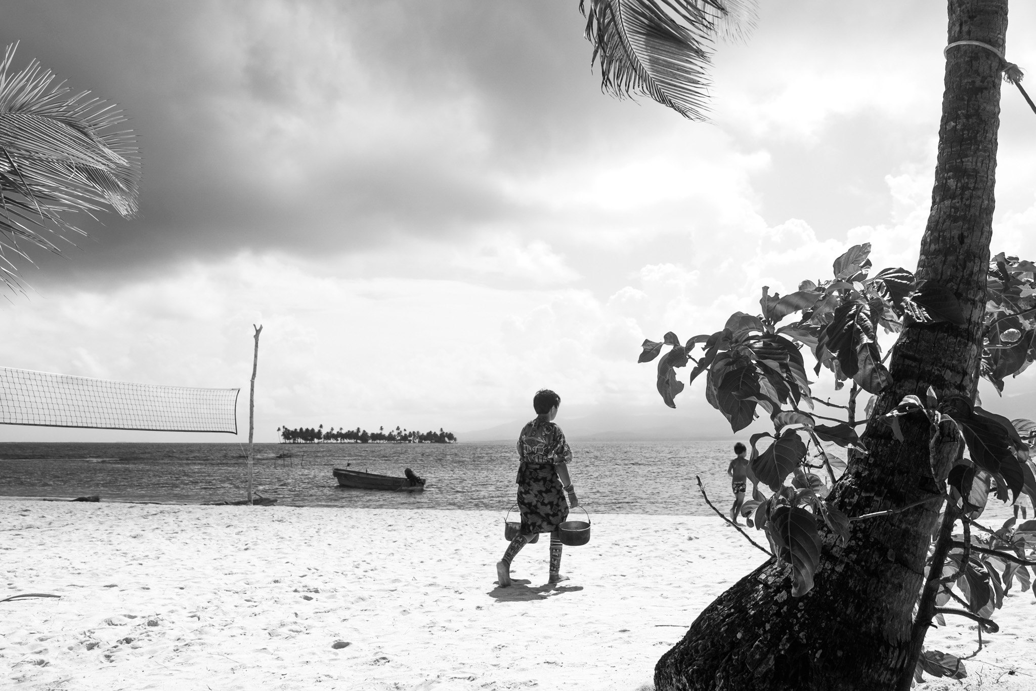 Mujer guna caminando por la playa con cubos, red de vóley y una isla al fondo